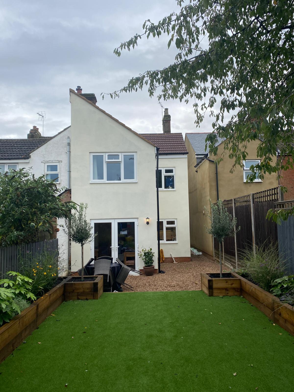 Rear garden with artificial lawn, sleeper planters and bark path behind a rendered house by DBD Builders Ltd